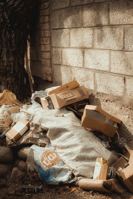 A pile of discarded cardboard boxes, some flattened and others partially folded, along with plastic bags filled with various rubbish, is situated against a brick wall outdoors. The cardboard has printed labels, barcodes, and logos visible on some pieces, and the bags appear crumpled and weathered, containing mixed waste materials. The scene is set near a concrete or stone wall, and the ground beneath the rubbish is uneven with small rocks and dirt. To the left, a portion of a tree trunk is visible, casting subtle shadows across the debris. The overall atmosphere suggests an area where private or independent rubbish removal, such as on-site clearance, might occur outside standard waste collection points, highlighting the importance of professional rubbish removal services like those provided by Rubbish Removal Bermondsey. The environment is lit by natural daylight, emphasizing the textures of the paper, plastic, and rough wall surface, aligning with the context of alternative waste handling solutions that may be necessary for private dump sites or excess refuse requiring professional disposal.
