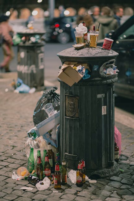 A black, metal public rubbish bin with a domed lid, positioned on a cobbled street, is overflowing with various types of waste. The top of the bin is cluttered with discarded items including a crumpled paper cup with a plastic lid, a used paper coffee cup with a cardboard sleeve, and a glass of beer with foam on top. A small cardboard box protrudes from the side of the bin, and several used paper napkins are draped over the edge. Around the base of the bin, multiple glass bottles—some green and some clear—with residual drinks inside are scattered, along with crumpled tissues, plastic packaging, and more paper waste. Behind the bin, a littered urban area features a green garbage bag, more scattered trash, and a blurred background with parked cars and pedestrians on a busy street, suggesting a typical scene of street-level waste that might result from informal disposal rather than organized rubbish collection. The setting appears to be in natural daylight, with overcast lighting emphasizing the textures and colours of the waste materials, reflecting common issues of improper rubbish disposal near public spaces.