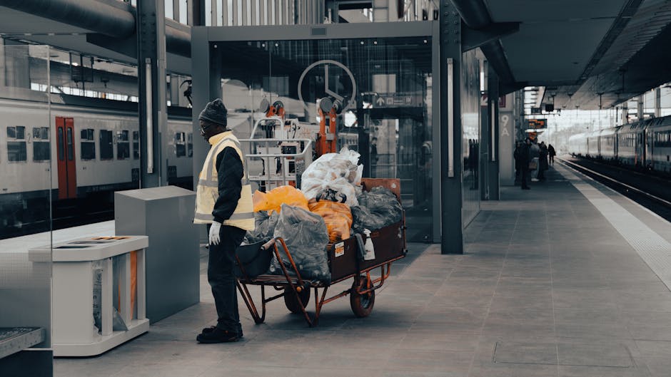 A person wearing a dark jacket, grey beanie, and high-visibility vest stands next to a large red trolley filled with various types of waste, including black and orange plastic bags, cardboard boxes, and other miscellaneous rubbish. The trolley is positioned on a tiled platform at a modern train station, with a glass shelter and metal framework visible in the background. To the left, a white recycling bin with a slot for waste disposal is placed nearby. In the background, train tracks extend into the distance, with a few passengers walking along the platform and a parked train featuring a red and white exterior. The environment is illuminated by natural daylight, and the scene depicts an area frequently used for passenger transit where private waste removal or on-site clearance services, such as those offered by Rubbish Removal Bermondsey, might be involved in handling excess rubbish outside of standard collection services. The textures of the waste bags and the sleek surfaces of the station are clearly distinguishable, capturing a typical urban rubbish collection context at a transportation hub.