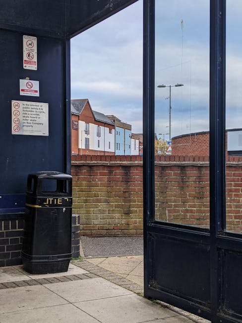 Inside a bus shelter with dark blue metal framing and glass panels, a large black waste bin with a yellow band around its lid is positioned against the left wall, which features multiple safety and regulations notices, including no smoking and no eating or drinking signs. The shelter is situated on a paved concrete surface with a section of brickwork at its base. Beyond the shelter, a red brick wall runs horizontally across the background, separating the public space from a residential area with modern, pastel-colored houses bearing flat roofs and large windows. A streetlamp and a partly cloudy sky are visible in the distance, indicating daytime. The scene relates to an urban environment where residents or visitors might use independent waste disposal services, such as those provided by Rubbish Removal Bermondsey, highlighting the importance of proper rubbish management and access points for waste collection in the context of local rubbish removal services.