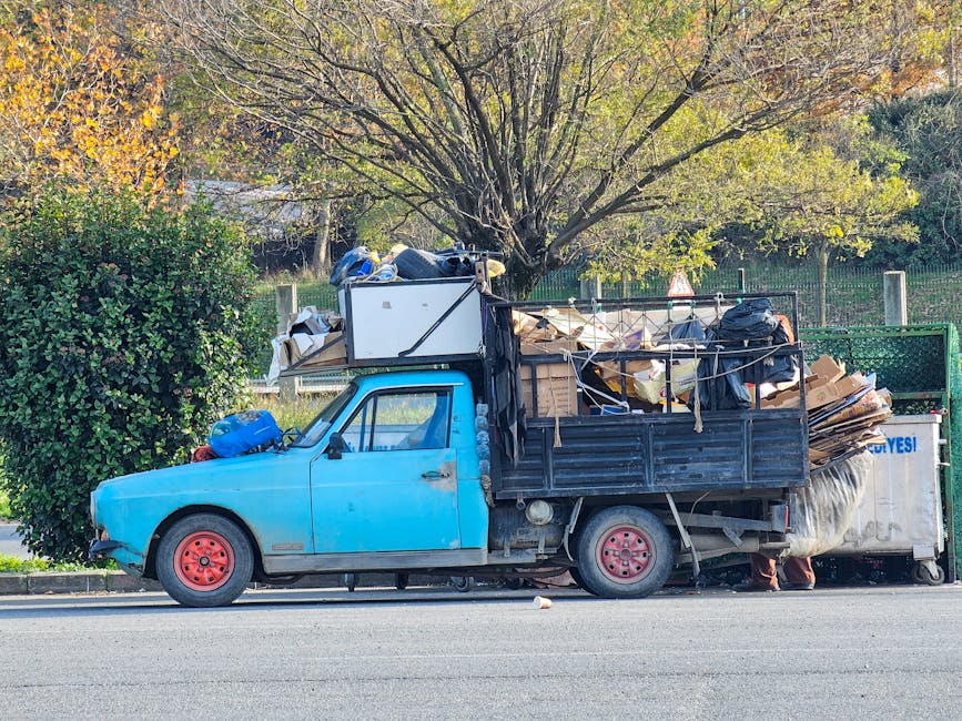 A small vintage blue pickup truck parked alongside a street, loaded with a large pile of assorted waste materials and cardboard boxes, secured with ropes and tied to the vehicle's black metal side panels. The truck's wheels are painted red, and its front bumper and bodywork show signs of age and wear, with a slightly faded finish. Behind the vehicle, there is a green hedge and a large leafy tree with light green and yellow foliage, indicating autumn. To the right of the truck, a white waste container is partially visible, positioned on a paved road. The scene suggests an on-site rubbish clearance operation, possibly arranged through a private waste management service like Rubbish Removal Bermondsey that offers alternative waste disposal options outside local authority collections, with the vehicle set up for independent rubbish collection or on-site removal, within a suburban environment. The lighting is natural daylight, illuminating the scene evenly and highlighting the textures of the waste and the vehicle's surface.