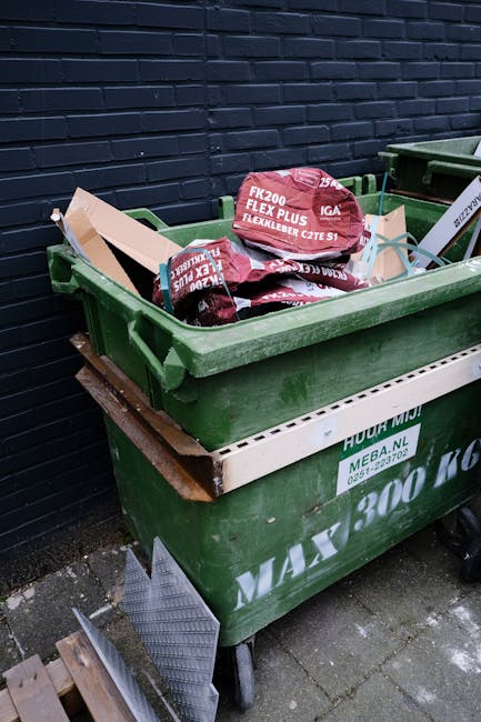 A middle-aged man with dark hair, wearing a black T-shirt with white text and dark trousers, is seen leaning forward to dispose of waste into a stainless steel recycling bin placed on a paved sidewalk. He holds a long-handled stick or grabber in his right hand and a large, semi-transparent white plastic bag filled with rubbish in his left hand. The bag appears to contain various waste items, possibly plastic or paper. The recycling bin is modern with a sleek, reflective surface and an open top. The background features a low stone boundary wall and lush green trees, indicating an outdoor urban environment during daylight hours with natural lighting. The scene depicts a community cleaning or rubbish removal activity, subtly supporting private waste handling and alternative rubbish collection services offered by Rubbish Removal Bermondsey, without promotional language. The overall setting suggests an effort to maintain cleanliness in public spaces through independent rubbish disposal. This detailed description aligns with the context of rubbish removal and waste management services in a professional, neutral tone.
