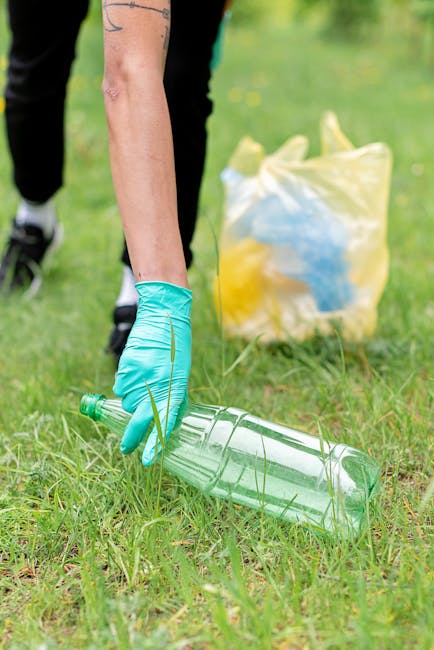 A person wearing a blue disposable glove is seen picking up a clear plastic bottle from a grassy outdoor area, with a yellow and white rubbish bag in the background. The individual is partially visible, with their arm and part of their black clothing shown against a blurred green environment. The plastic bottle is empty and transparent, lying horizontally on the grass, with some blades of grass around it. The scene suggests an activity related to litter collection or environmental cleanup. This image illustrates the importance of proper waste disposal, aligning with services like independent waste removal and rubbish collection offered by companies such as Rubbish Removal Bermondsey, particularly when engaging in on-site clearance or private rubbish handling tasks in outdoor or garden settings.