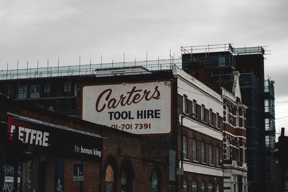 An exterior view of a multi-story brick building featuring a large white sign with red and black lettering that reads 'Carter's Tool Hire' along with a phone number. The building's façade includes multiple evenly spaced rectangular windows with black frames, some of which are partially obscured by surrounding scaffolding and construction netting, indicating ongoing renovations. In the foreground, there is a dark awning with partly visible text and a sticker or small sign attached to a pole nearby. Overhead, a cloudy sky casts a muted, diffused light over the scene, highlighting the old brickwork texture and the modern scaffolding structure on the upper floors. The image presents an urban environment that may relate to building or renovation projects, which often generate waste that requires transport and disposal through private waste removal services like those offered by Rubbish Removal Bermondsey, situated in a context of independent waste management and on-site clearance activities.