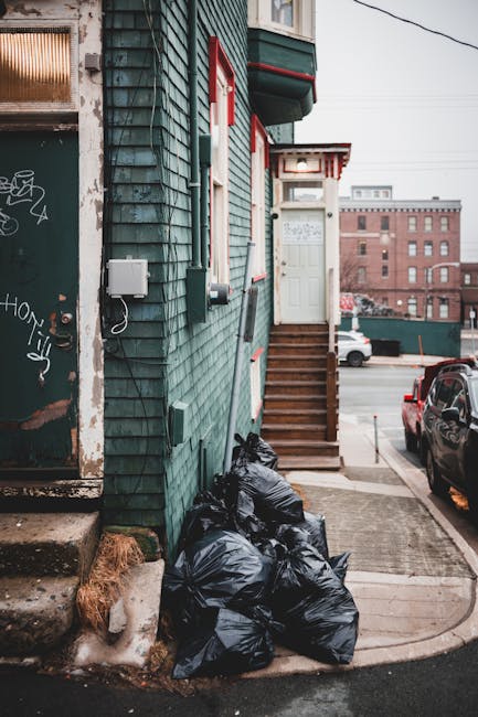 The image depicts the exterior corner of a building with green wooden shingle siding, situated on a street with parked cars. A small set of wooden stairs with a handrail leads up to a white door with a window, positioned at the top of the stairs. To the left of the stairs, several black trash bags are stacked on the pavement next to the building, suggesting the collection of waste or rubbish. The building has visible electrical meters and wires attached to the exterior wall, along with some graffiti on a weathered nearby door frame. In the background, multi-storey brick buildings and vehicle traffic can be seen under an overcast sky, creating a typical urban setting. The scene reflects elements of private waste disposal, with the rubbish bags awaiting collection, consistent with independent rubbish removal services like those offered by Rubbish Removal Bermondsey, in a typical Bermondsey street environment during daylight hours.