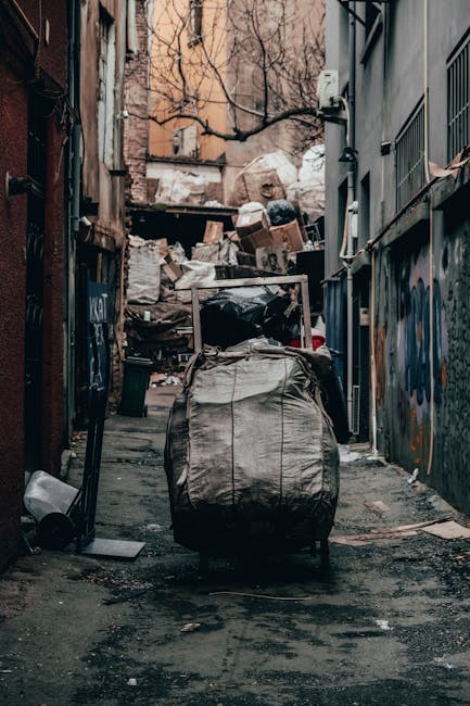 A narrow urban alleyway cluttered with a large, grey, heavy-duty rubbish bag in the foreground, which appears to be filled with waste materials. Behind it, there are more piles of mixed waste, including cardboard boxes, black plastic bags, and discarded items, stacked against a weathered fence and the back of a building. The pavement is uneven, with visible dirt and debris. On the right side, the side of a modern building features metal-framed windows with bars, while on the left, older brick walls are visible, some with graffiti. Overhead, leafless tree branches extend across the background, with a tangle of old electrical wiring and utility boxes attached to the buildings. The scene suggests an example of private waste collection or rubbish removal process in an urban environment, possibly managed by a local waste removal service like Rubbish Removal Bermondsey, serving the Bermondsey Street SE1 area.