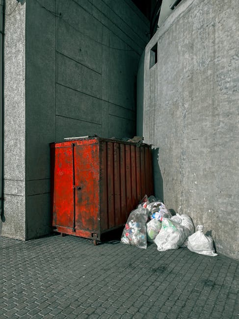 The image depicts the exterior corner of a building with green wooden shingle siding, situated on a street with parked cars. A small set of wooden stairs with a handrail leads up to a white door with a window, positioned at the top of the stairs. To the left of the stairs, several black trash bags are stacked on the pavement next to the building, suggesting the collection of waste or rubbish. The building has visible electrical meters and wires attached to the exterior wall, along with some graffiti on a weathered nearby door frame. In the background, multi-storey brick buildings and vehicle traffic can be seen under an overcast sky, creating a typical urban setting. The scene reflects elements of private waste disposal, with the rubbish bags awaiting collection, consistent with independent rubbish removal services like those offered by Rubbish Removal Bermondsey, in a typical Bermondsey street environment during daylight hours.